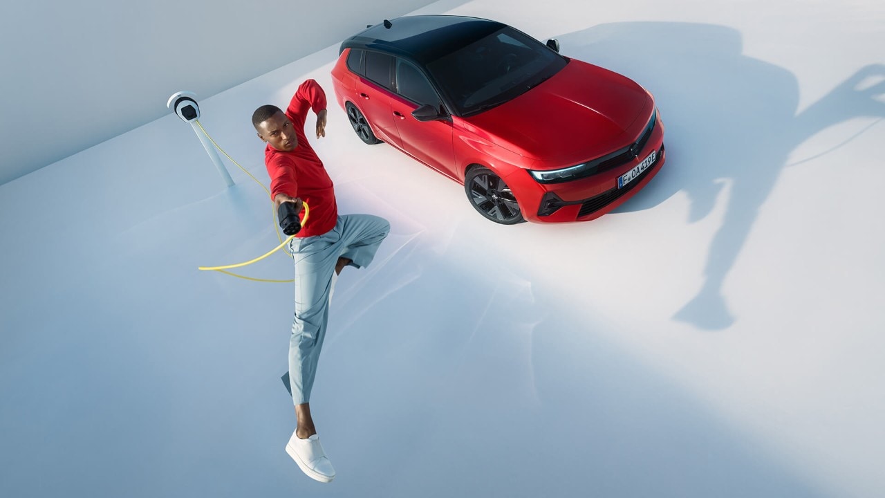 Hombre saltando en el aire con una estación de recarga de vehículos eléctricos y un Opel Astra Sports Tourer Electric de fondo.