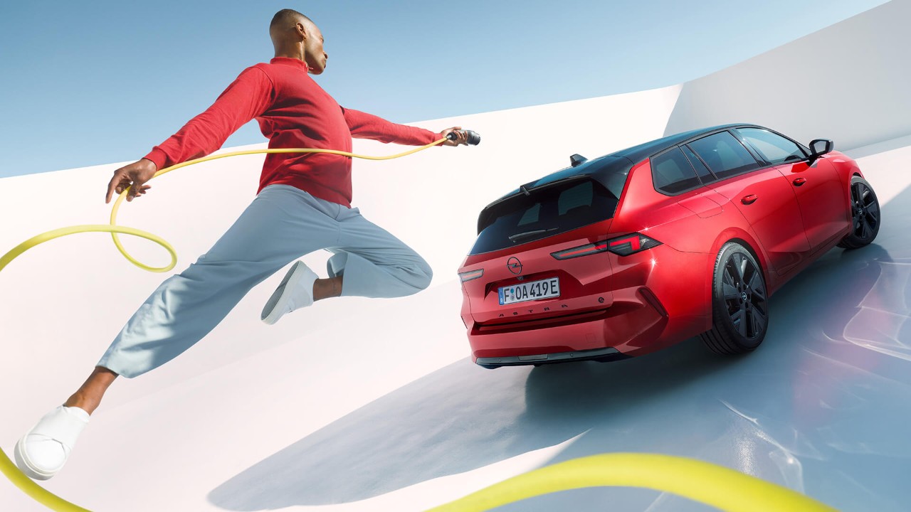 Hombre saltando en el aire con un cable de carga de un vehículo eléctrico con un Opel Astra Sports Tourer Electric de fondo.