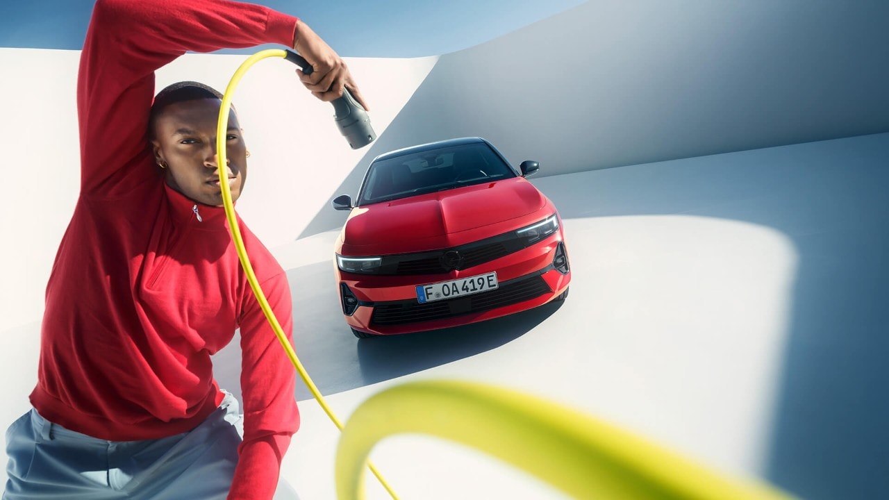 Hombre sosteniendo un cable de carga de un vehículo eléctrico delante de un Opel Astra Sports Tourer rojo con techo negro.