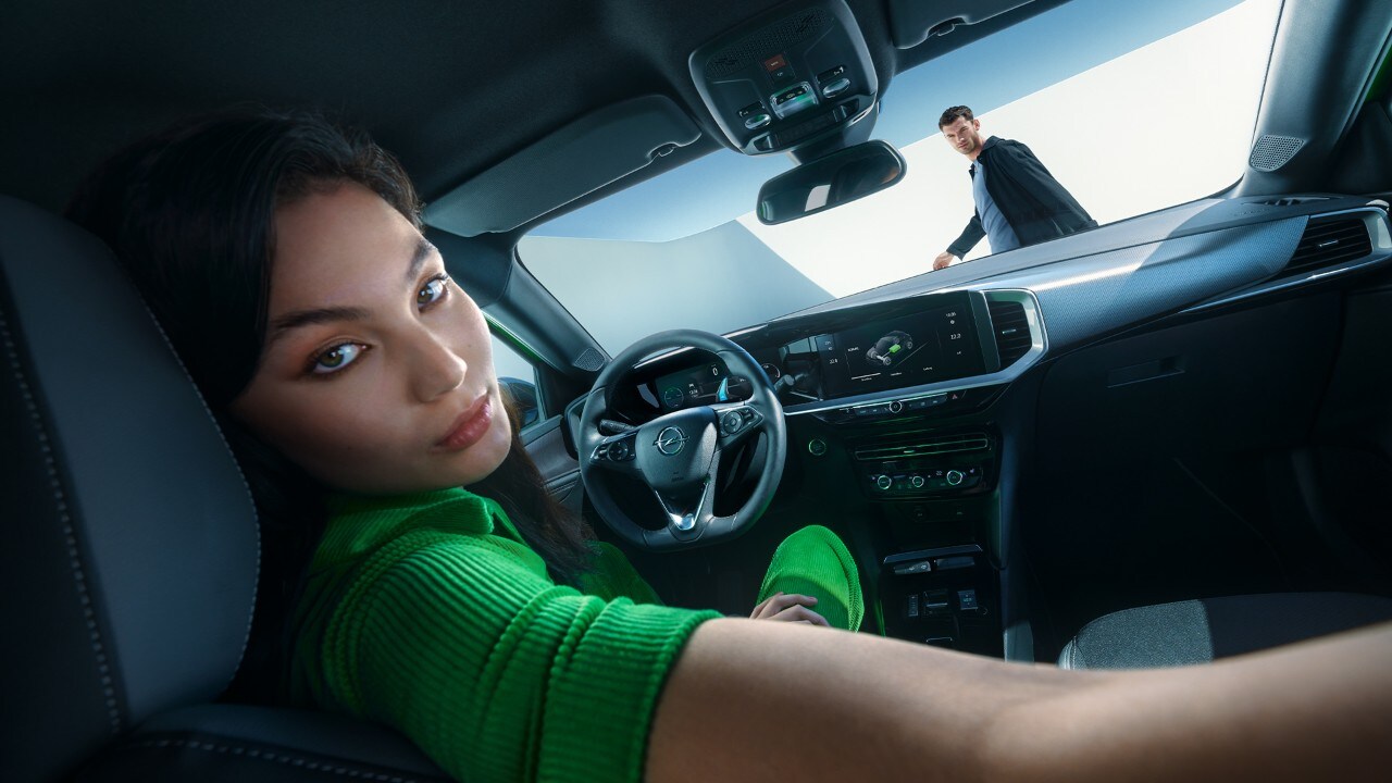 Mujer mirando hacia atrás mientras está en el asiento del conductor de un Opel Mokka Electric con un hombre caminando fuera