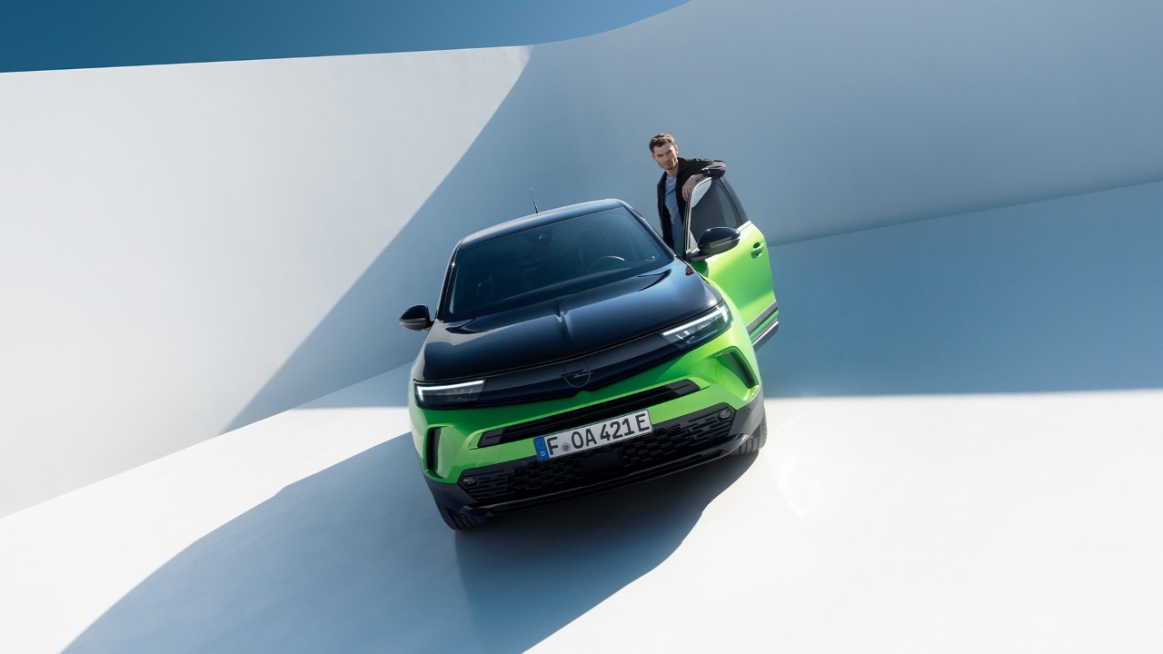 Vista frontal de un Opel Mokka Electric verde con aleaciones negras y techo con un hombre entrando por la puerta del conductor