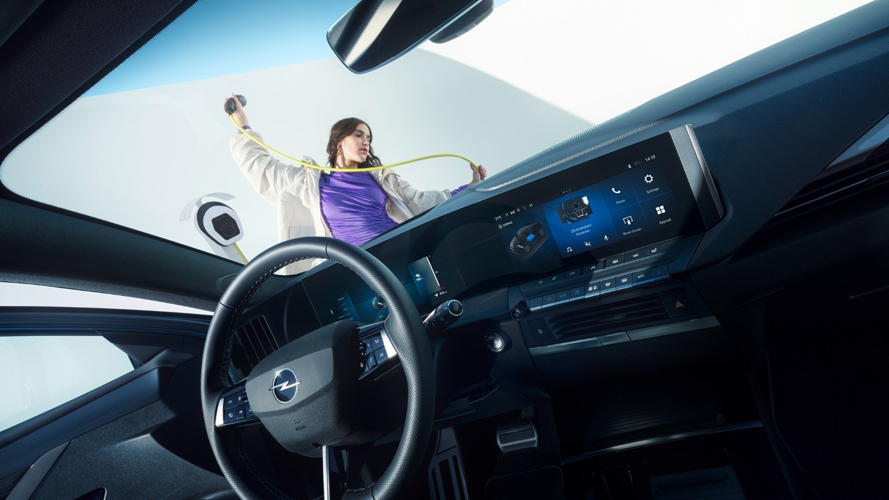 Vista interior de un Opel Astra Electric desde el asiento del copiloto, con una mujer con un cable de carga en el exterior.